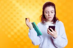 Una mujer joven con expresión confusa sostiene un smartphone, una tarjeta de crédito y un cuaderno con un resumen de oferta, ilustrando el desafío de cómo comparar ofertas de crédito.
